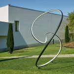 A black and white painted stainless steel loop sculpture sits on a lawn in front of a grey building.