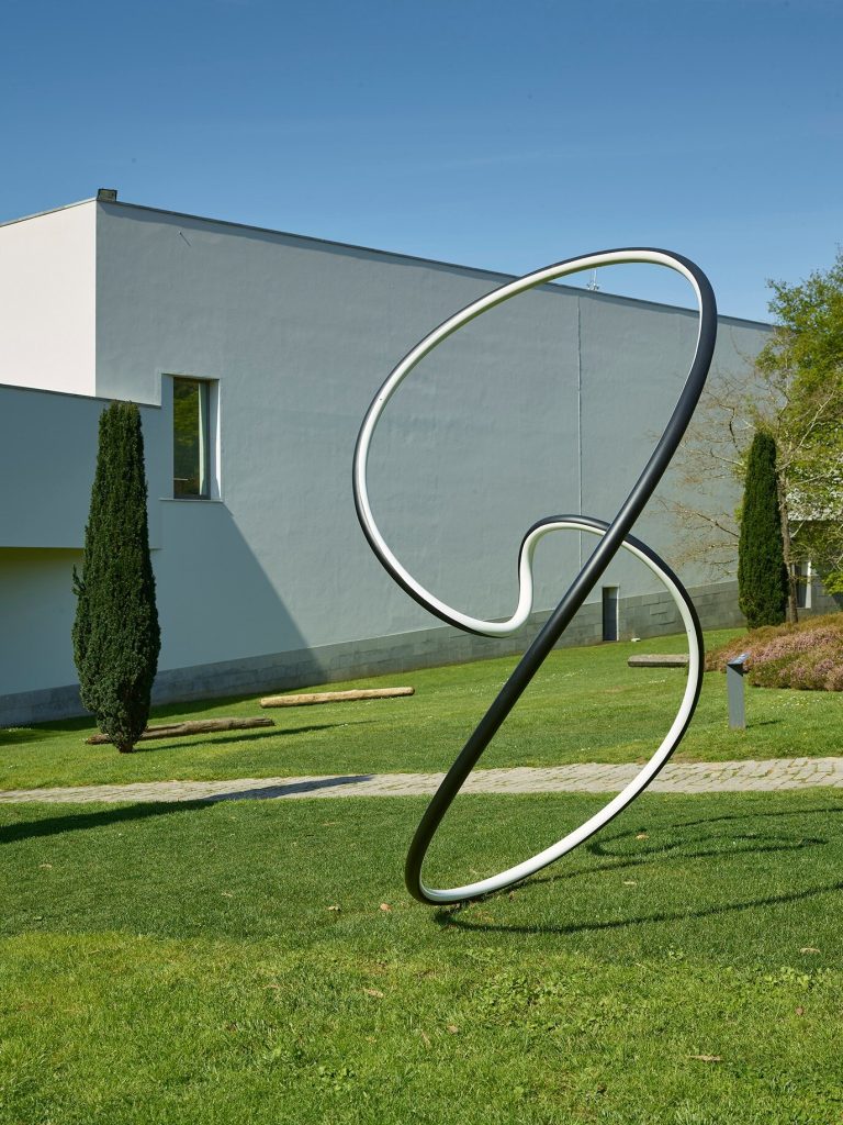 A black and white painted stainless steel loop sculpture sits on a lawn in front of a grey building.