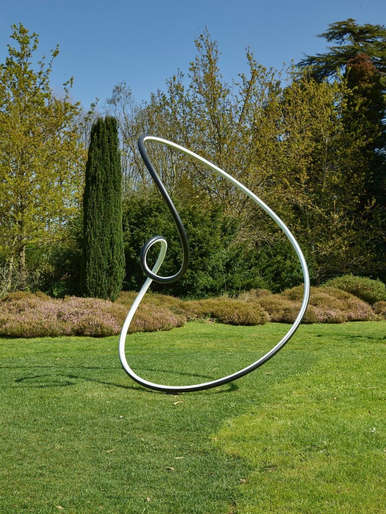 A black and white painted stainless steel sculpture sits on a lawn with trees in the background.