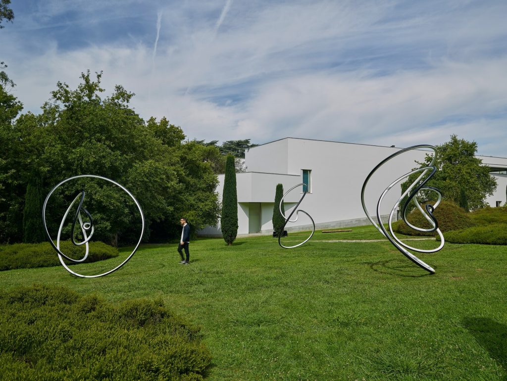 Three stainless steel sculptures with black and white paint sit on a grassy lawn near a white building under a cloudy sky.