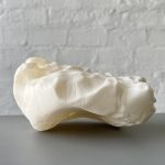 A white 3D printed resin object with a wavy surface sits on a gray table against a white brick wall.