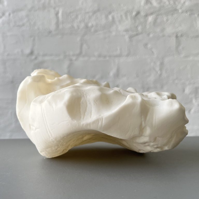 A white 3D printed resin object with a wavy surface sits on a gray table against a white brick wall.