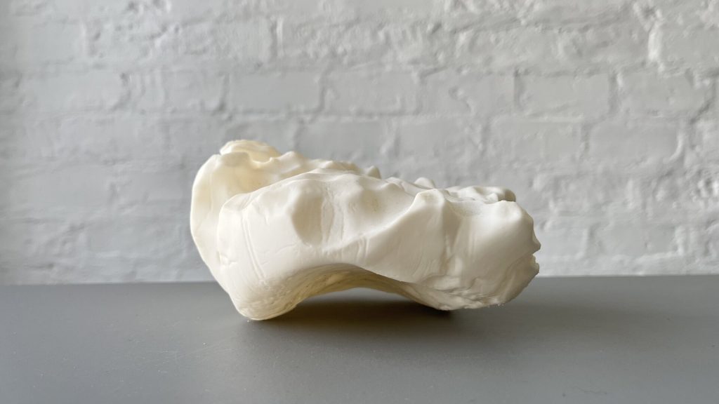 A white, irregularly shaped 3D-printed resin object sits on a gray surface against a white brick wall.