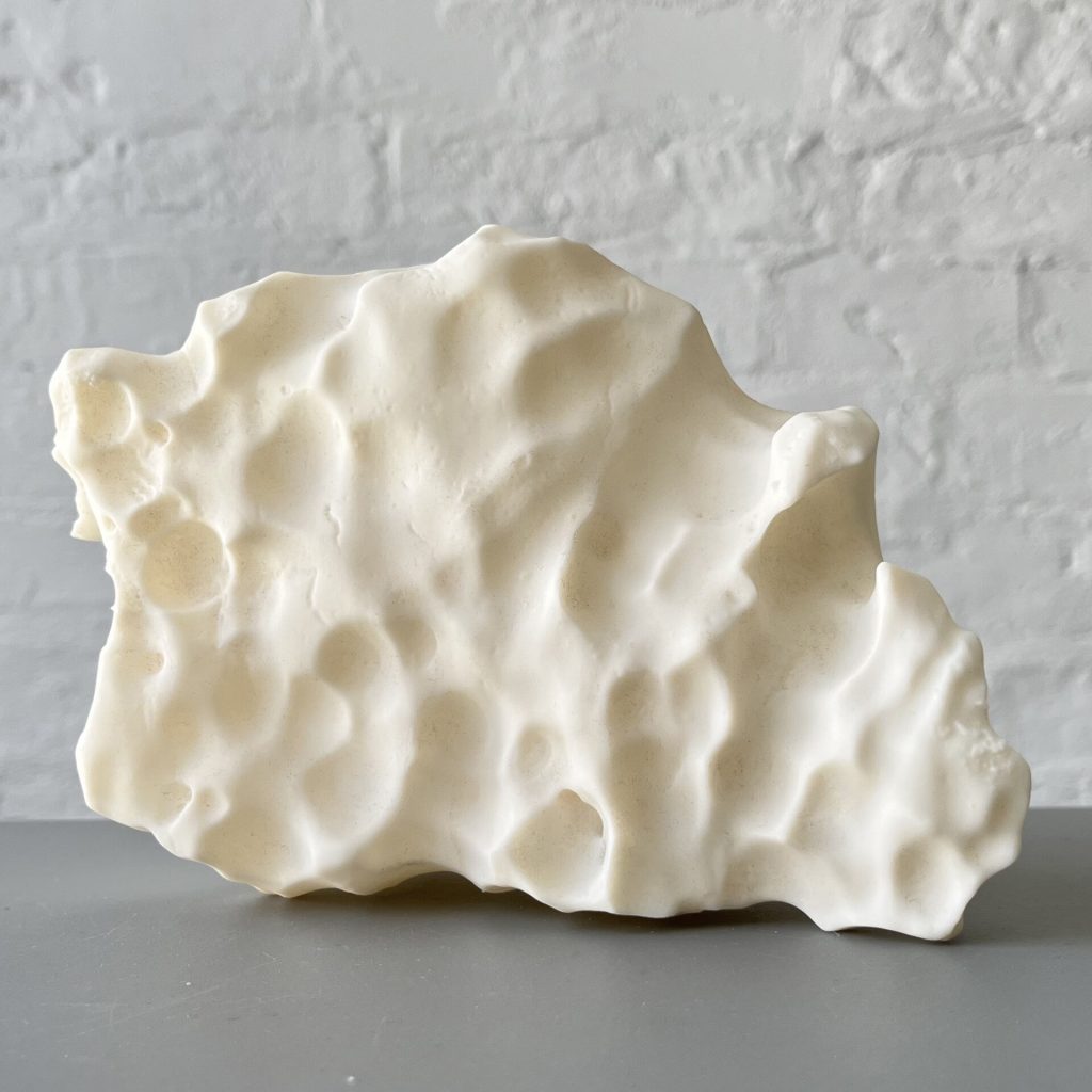 A 3D printed resin block with pitted, uneven surfaces sits on a gray shelf against a white brick wall.