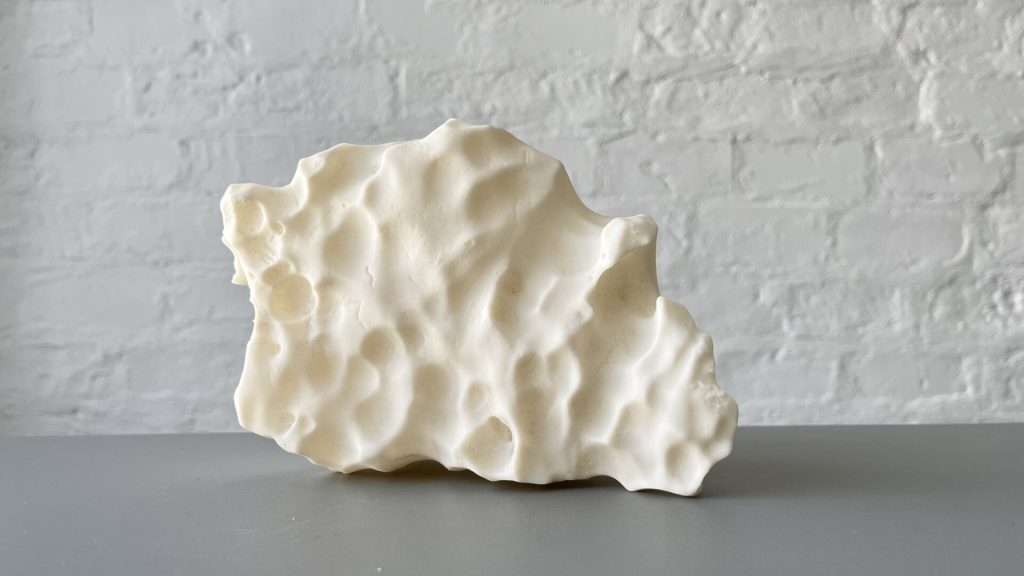 A white, irregularly shaped object made of 3D-printed resin sits on a gray surface against a white brick wall.