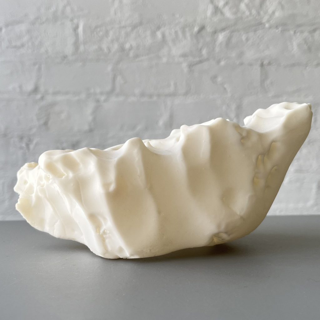 A white 3D printed resin object with a wavy, melting surface sits on a gray table against a white brick wall.