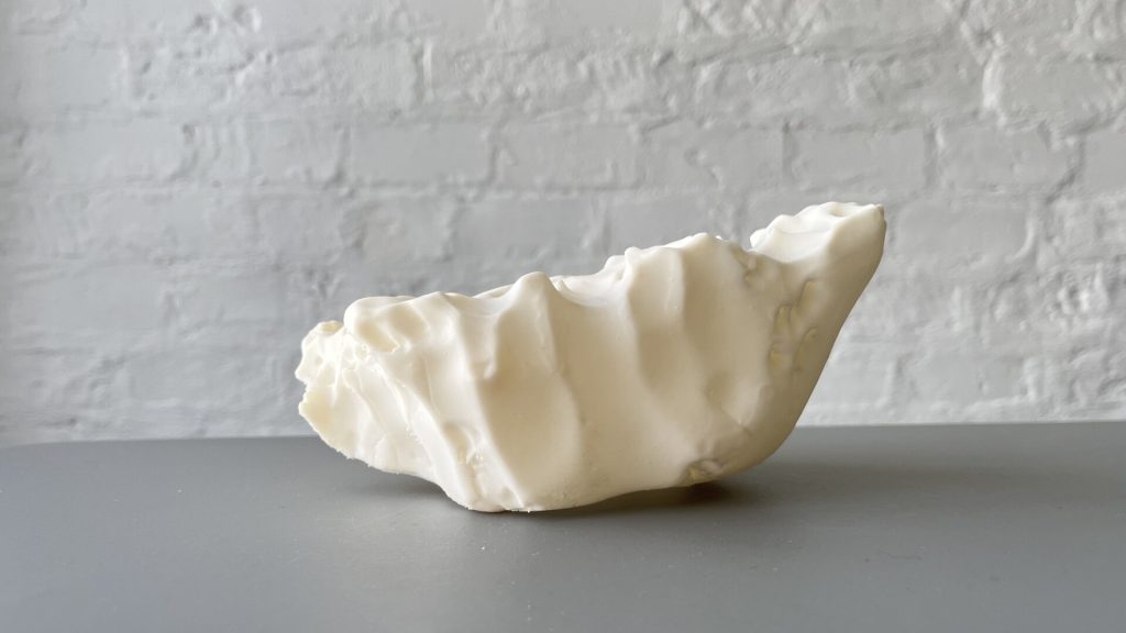 A white, irregularly shaped 3D-printed resin object sits on a gray surface against a white brick wall.