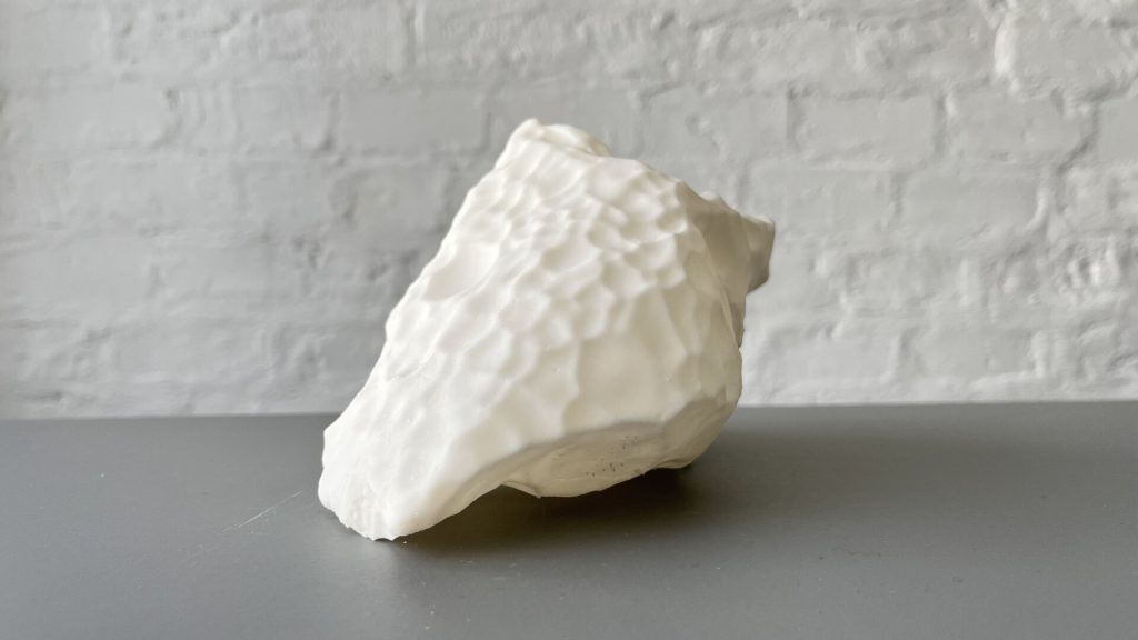 A white, textured 3D-printed resin object sits on a gray surface against a white brick wall.