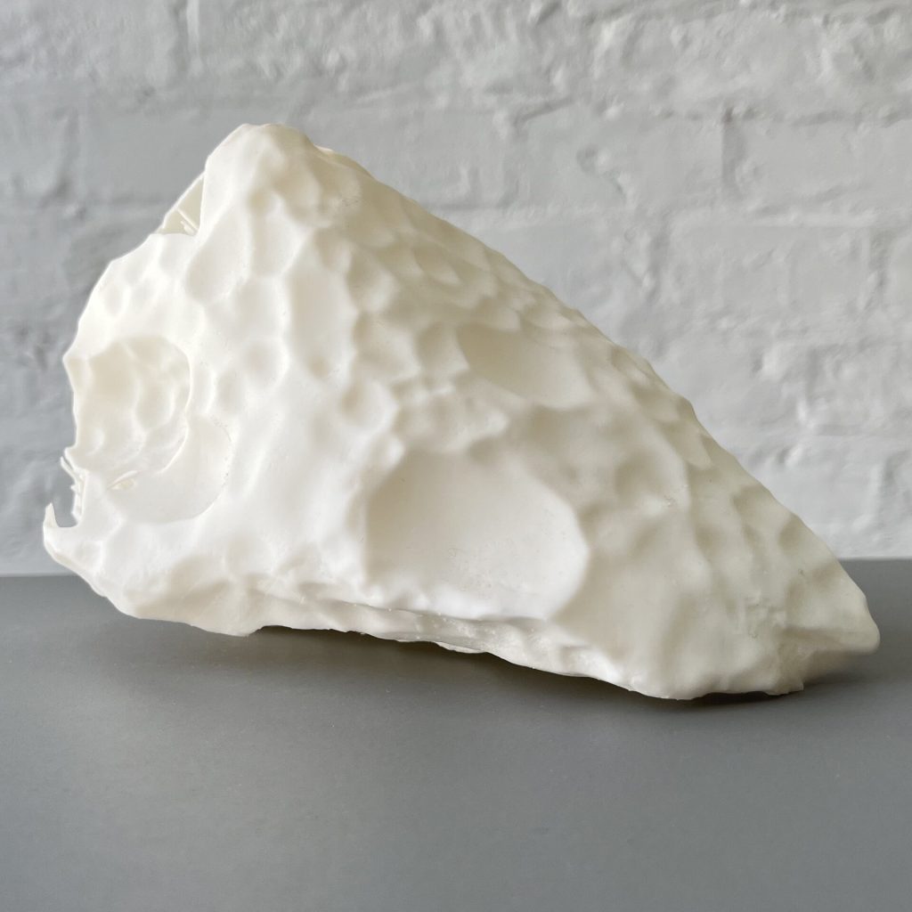 A white 3D printed resin block with a bumpy surface sits on a gray table against a white brick wall.