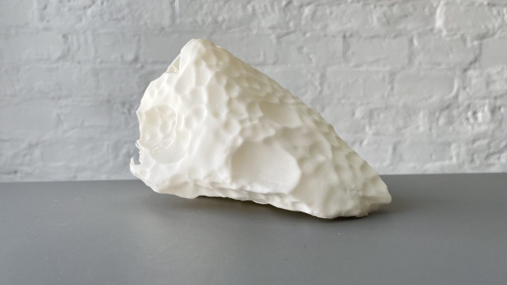 A white, textured 3D-printed resin object sits on a gray surface against a white brick wall.
