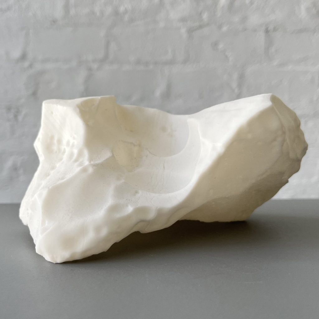 A white 3D printed resin object sits on a gray surface against a white brick wall.