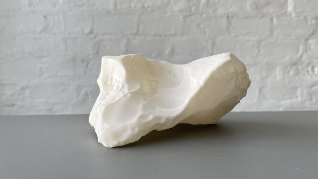 A white, irregularly shaped 3D-printed resin object sits on a gray surface against a white brick wall.