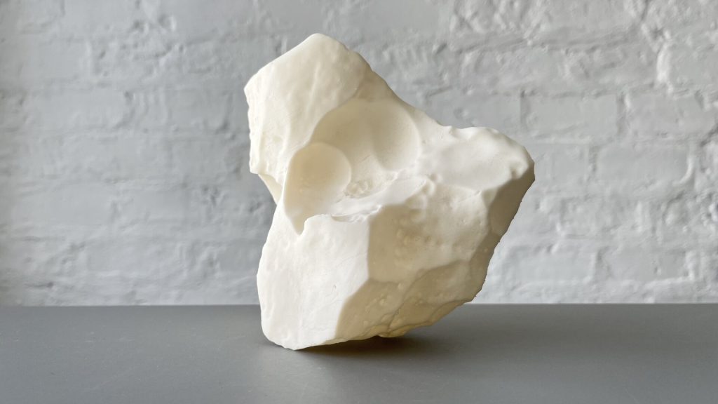 A white, irregularly shaped 3D-printed resin object sits on a gray surface against a white brick wall.