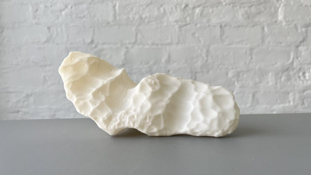 A white, irregularly shaped 3D-printed resin object sits on a gray surface against a white brick wall.