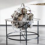 A stainless steel frame holds a glass sphere with mirrors and 3D prints on a metal table.