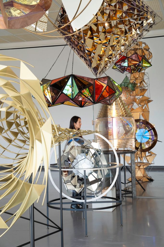 A woman stands inside a stainless steel and glass Kaleidosphere sculpture surrounded by hanging geometric light fixtures.