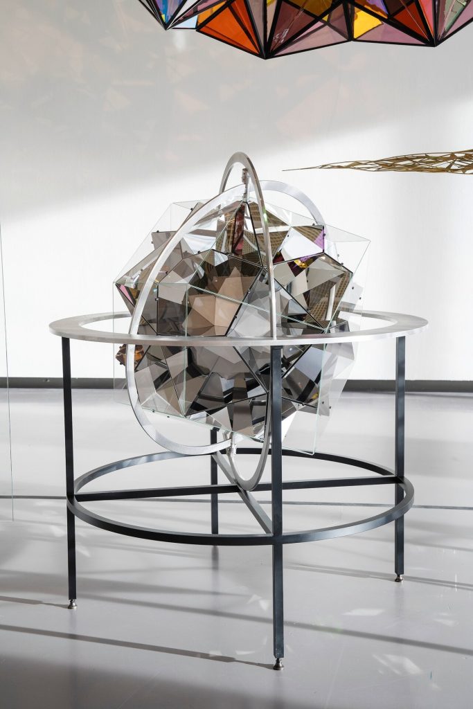 A stainless steel frame holds a mirrored, faceted sphere on a circular base in a gallery space.