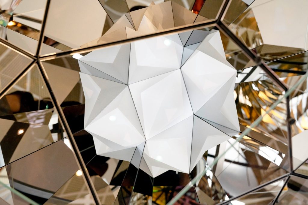 A white, geometrically faceted sphere hangs within a mirrored structure composed of stainless steel frames and reflective panels.