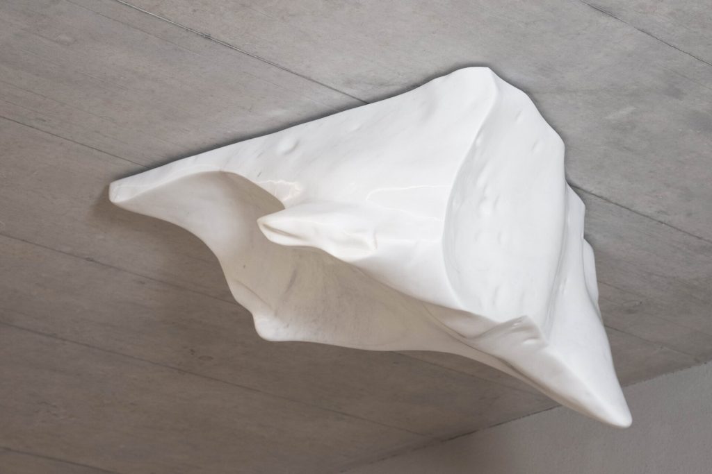 A white Carrara marble sculpture hangs from the ceiling in a minimalist gallery space.