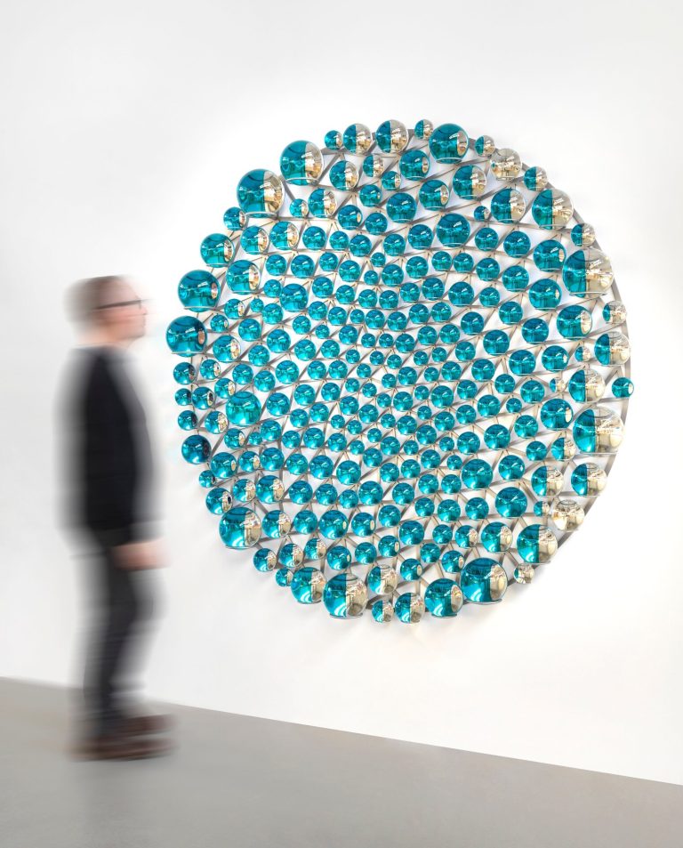 A circular wall installation of cyan and silver glass spheres hangs on a white wall.