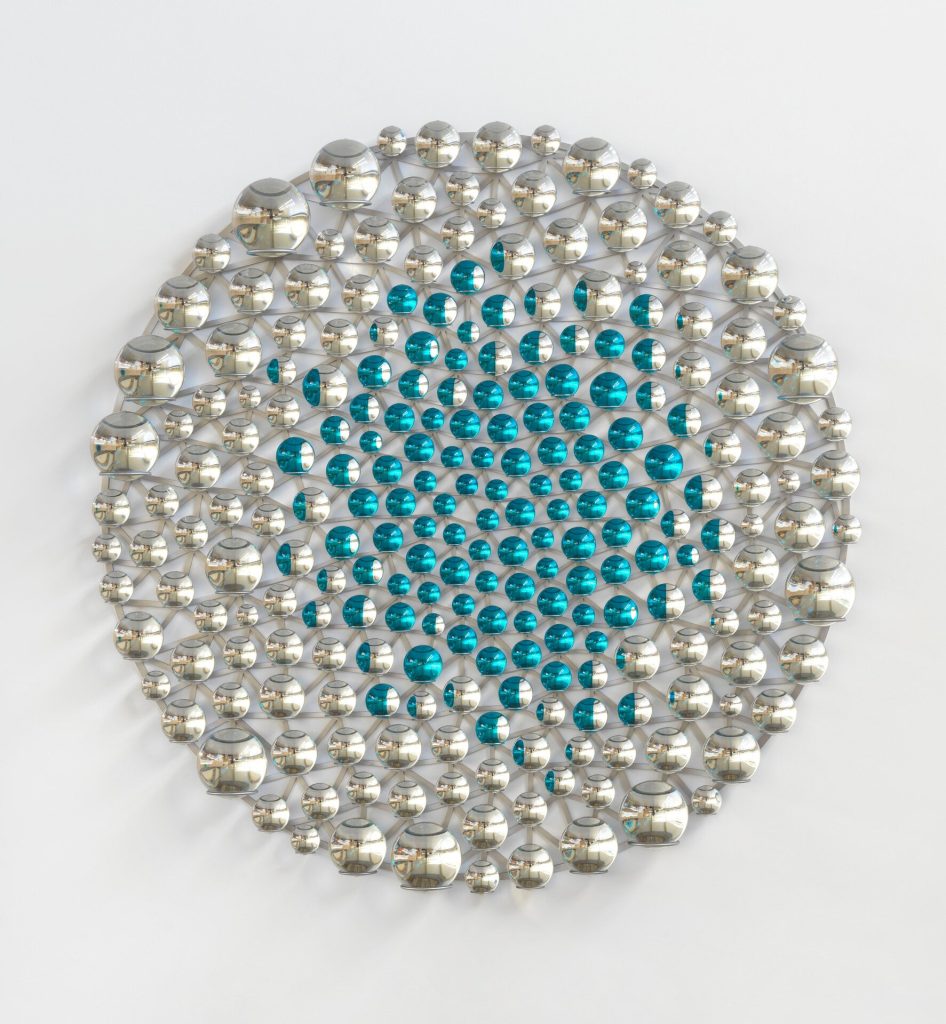 A circular stainless steel grid holds silvered glass spheres with cyan paint centers on a white wall.