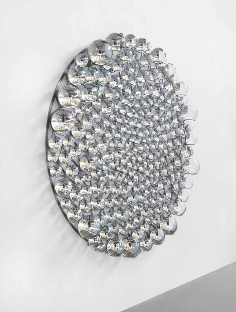 A stainless steel disc holds partially silvered glass spheres with cyan and black paint on a white wall.