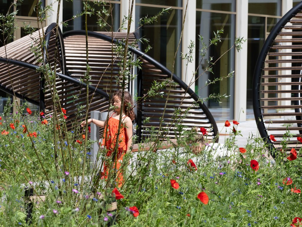 A girl in an orange dress stands among red poppies near a painted stainless steel and wood spiral structure.