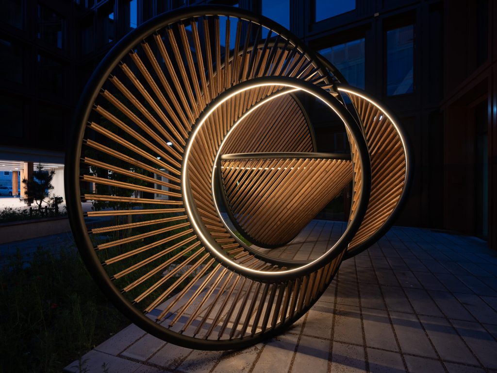 A painted stainless steel and wood sculpture with integrated lighting sits on a paved surface.