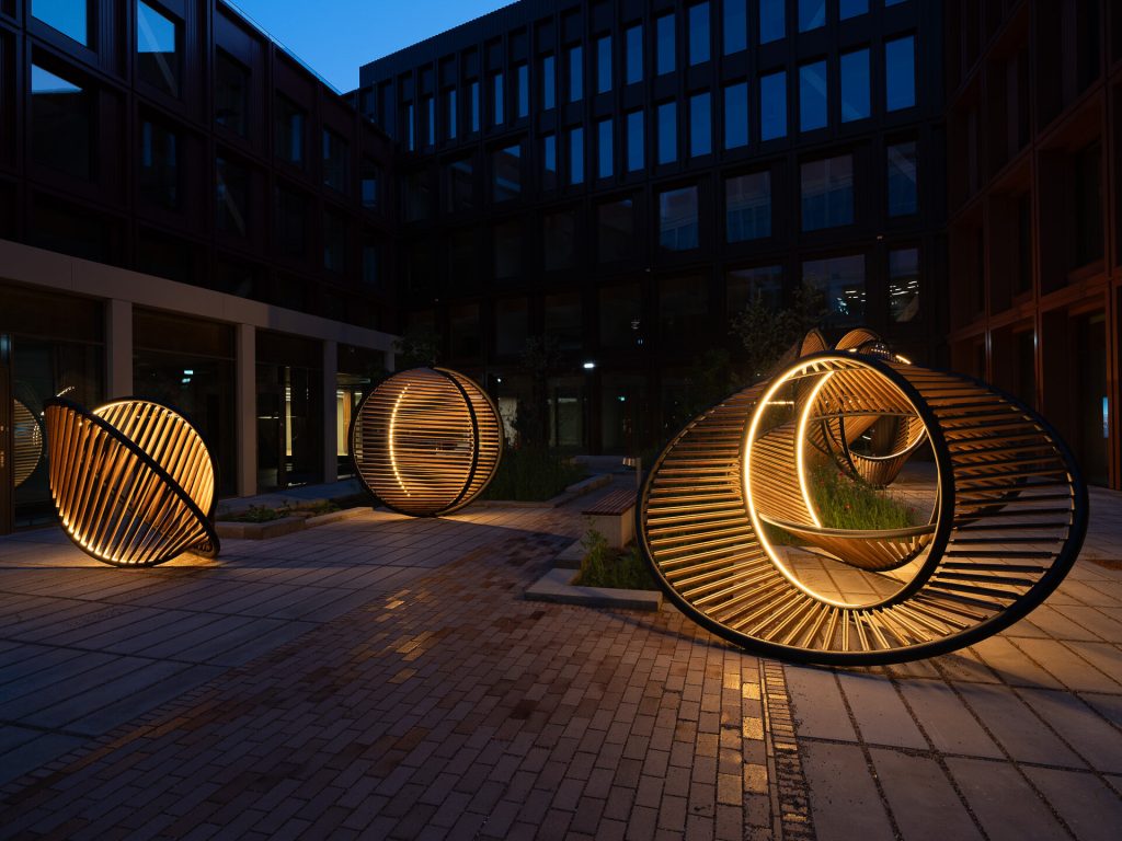 Three illuminated wooden sculptures with integrated lighting sit on a brick courtyard surrounded by modern buildings at dusk.