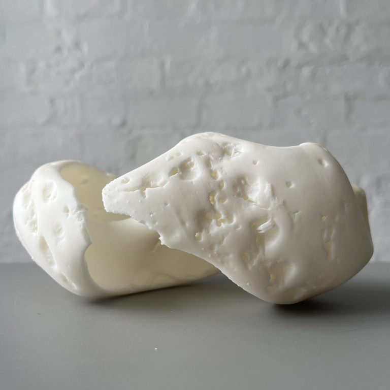 A white 3D printed resin object with porous holes sits on a gray surface against a white brick wall.