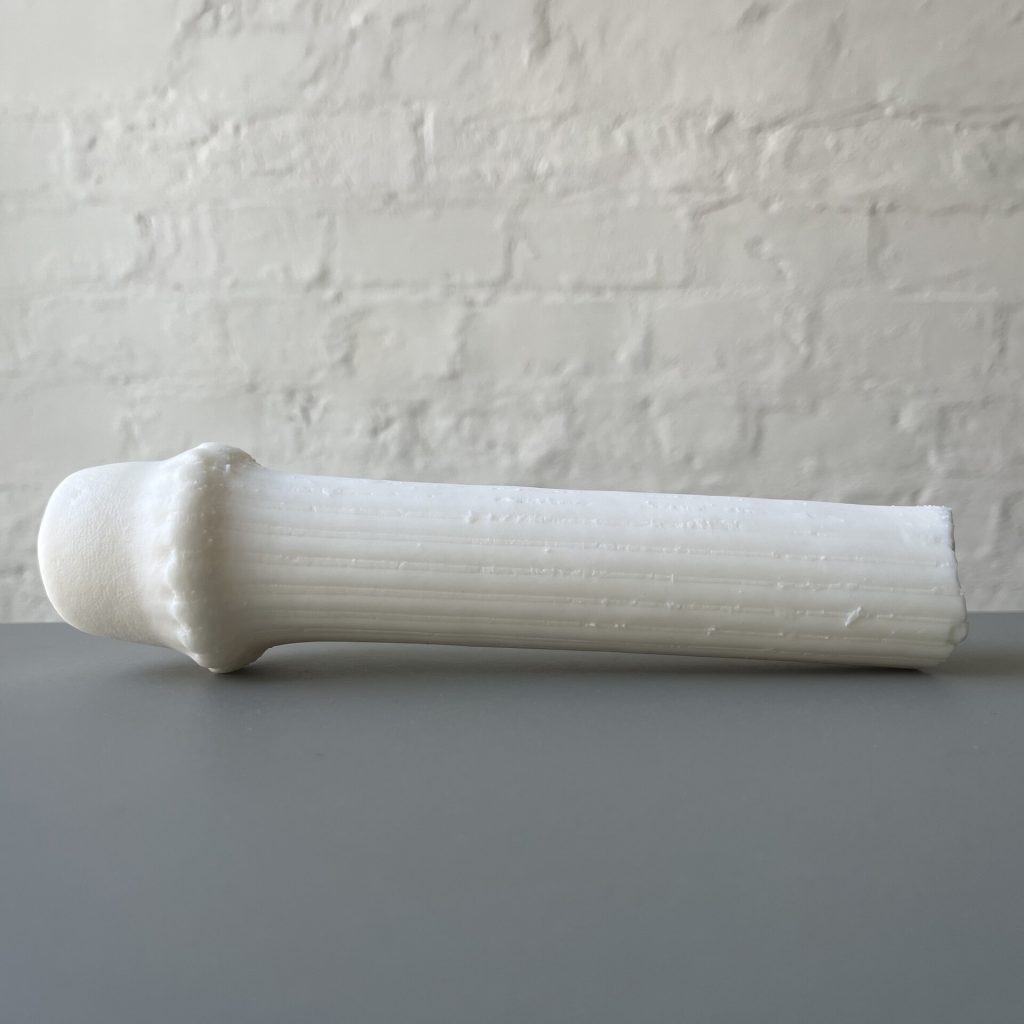 A white 3D printed resin cylinder with a rounded head sits on a gray surface against a white brick wall.