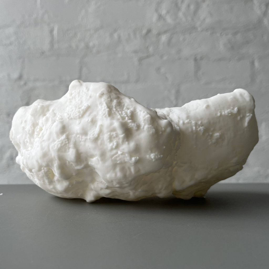 A white 3D printed resin object with a bumpy texture sits on a gray surface against a white brick wall.