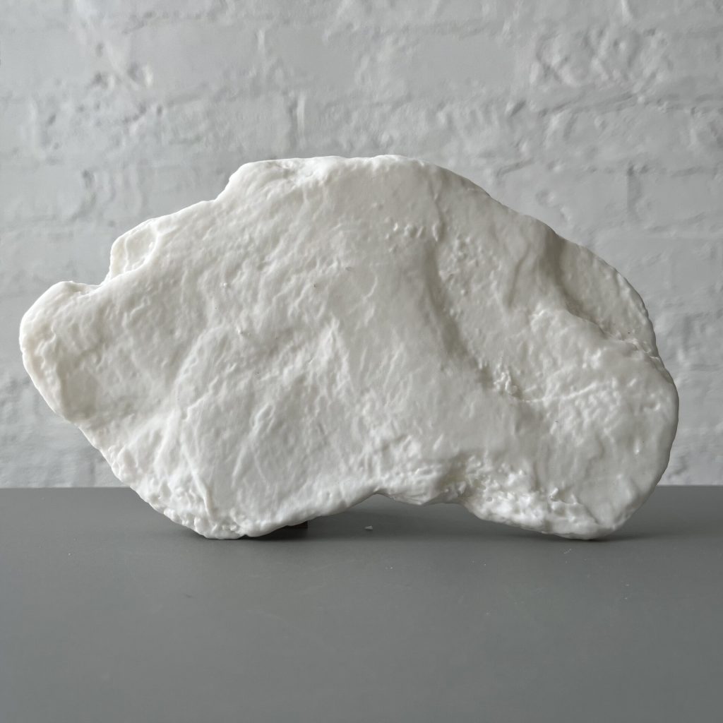 A white 3D printed resin object sits on a gray surface against a white brick wall.