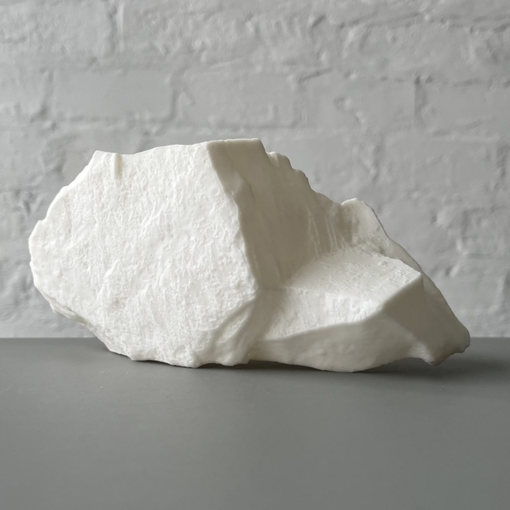 A white 3D printed resin object sits on a gray surface against a white brick wall.