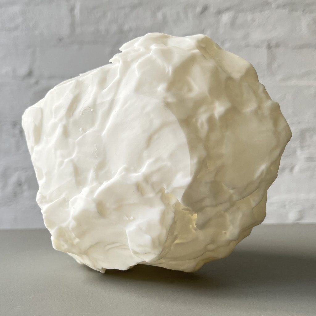 A white 3D printed resin object with a crinkled surface sits on a gray shelf against a white brick wall.