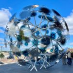 A segmented spherical sculpture with blue-tinted circular panels sits on a paved plaza under a blue sky.