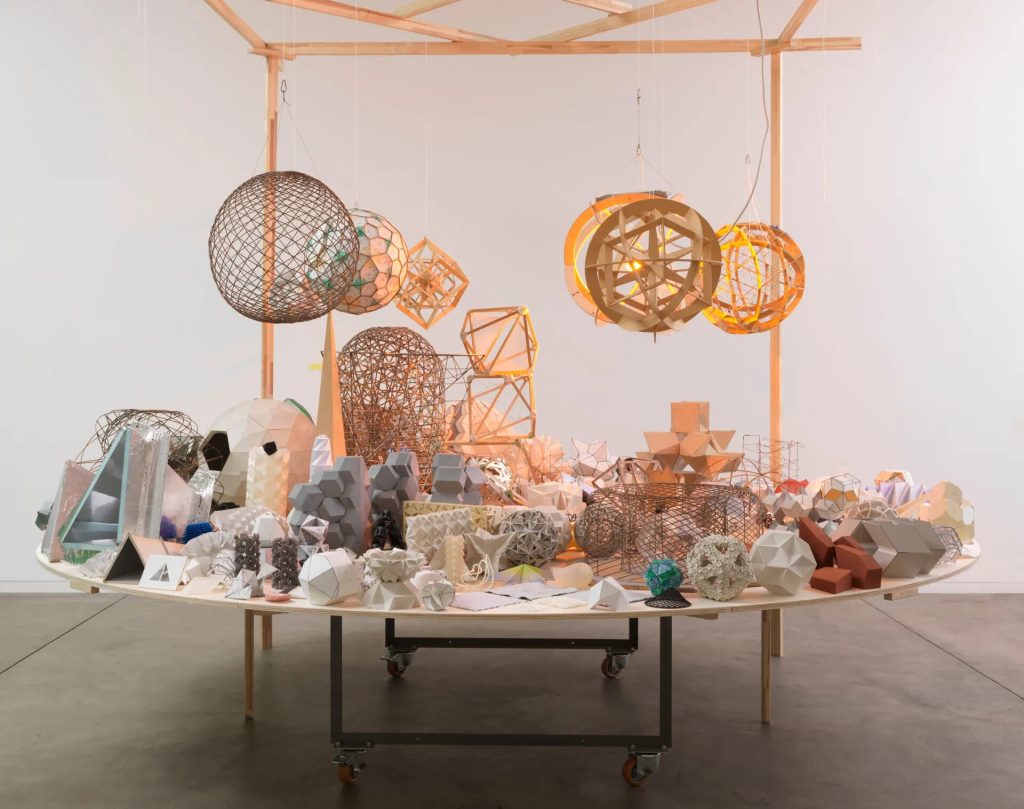 A wooden table displays mixed-media geometric models under hanging structures in an art installation.