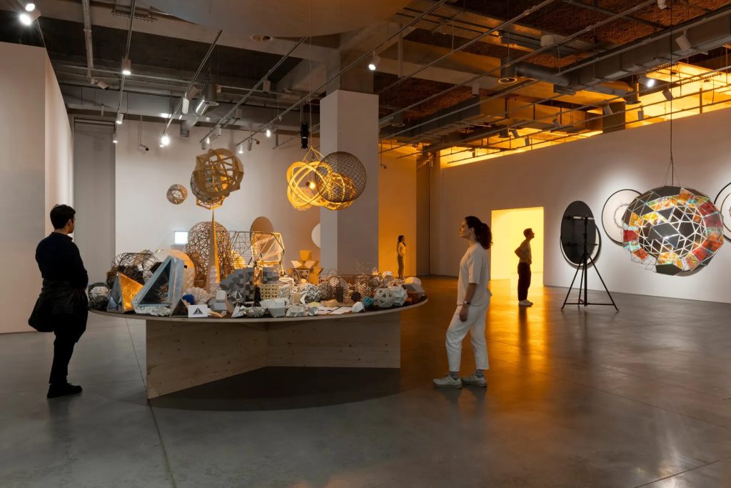 A wooden table displays mixed-media geometric models in an art gallery with hanging sculptures and visitors observing the exhibit.