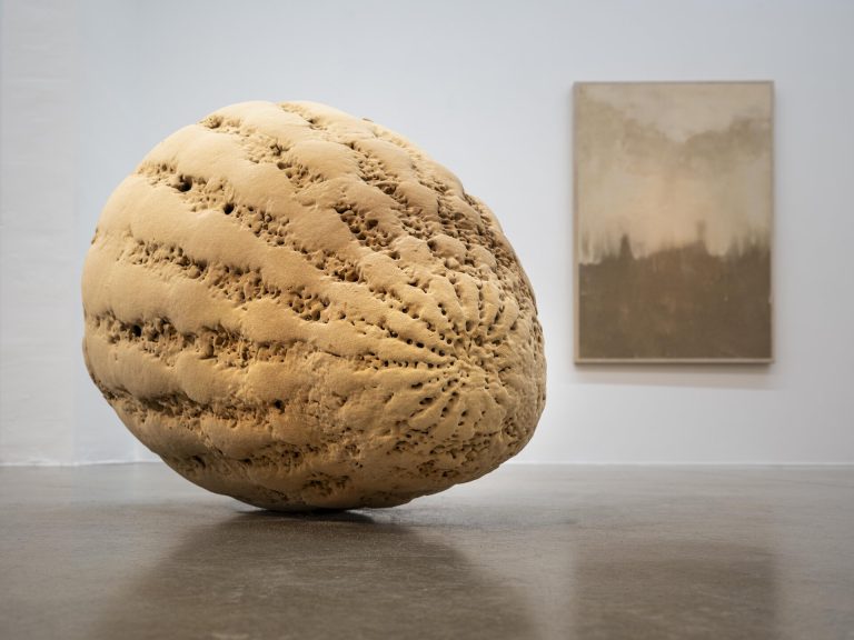 A large, textured, sand-like sculpture sits on a concrete floor with a muted painting hanging on the wall behind it.