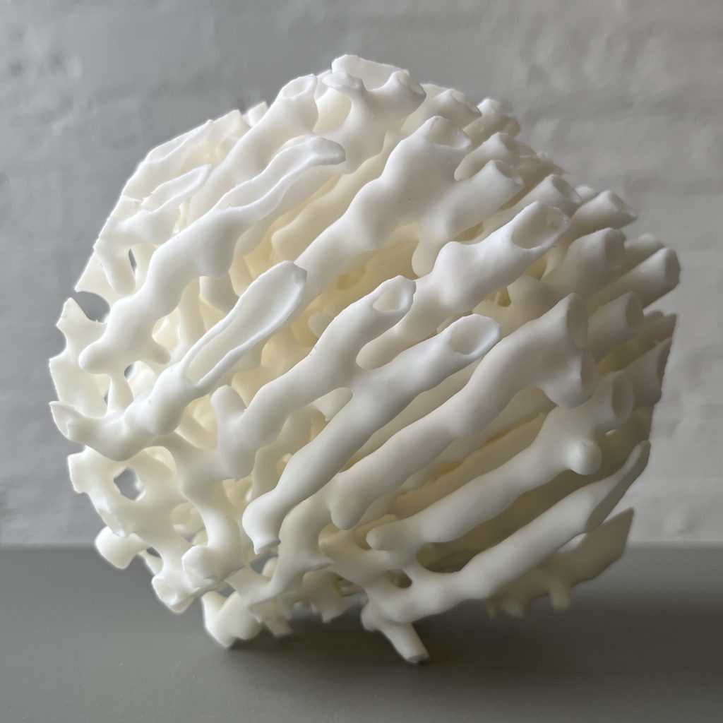 A white 3D printed resin sphere with porous, branching structures sits on a gray surface.