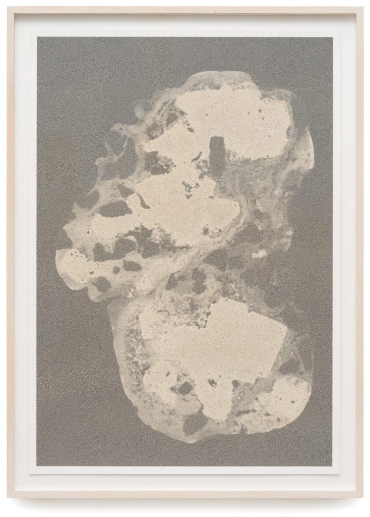A framed screen print displays sand particles from Senegal embedded in ink on paper, forming an abstract shape against a textured gray background.