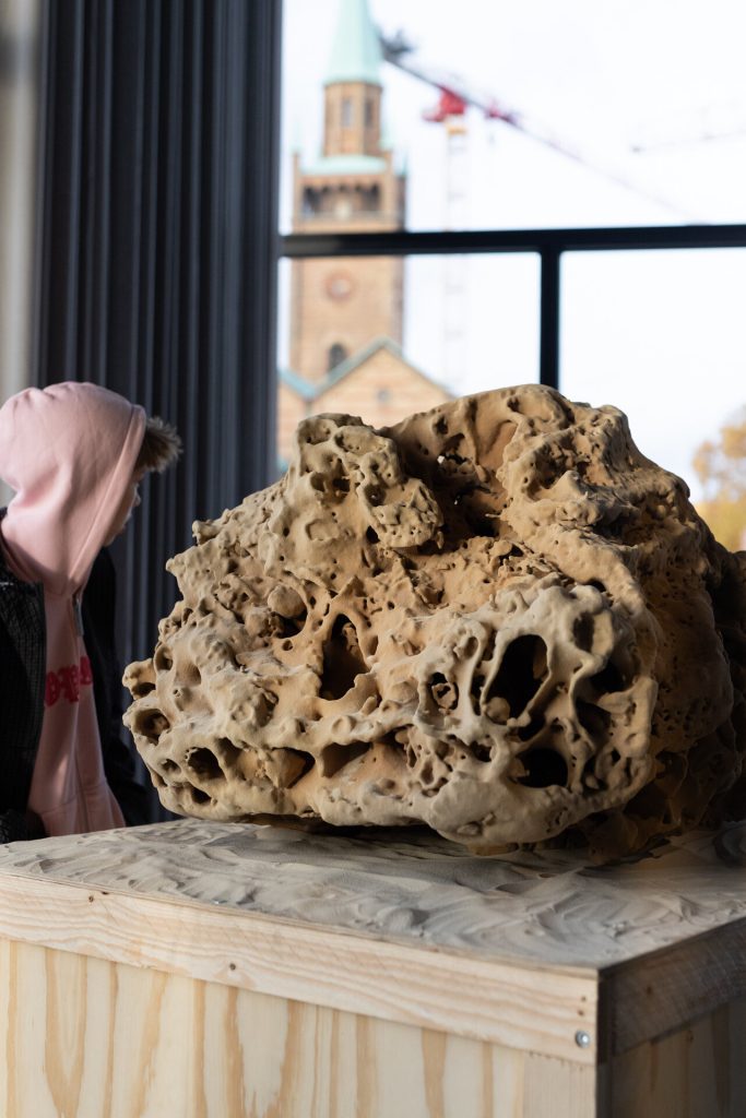 A porous 3D-printed sand sculpture sits on a wooden crate while a person in a pink hoodie looks on.