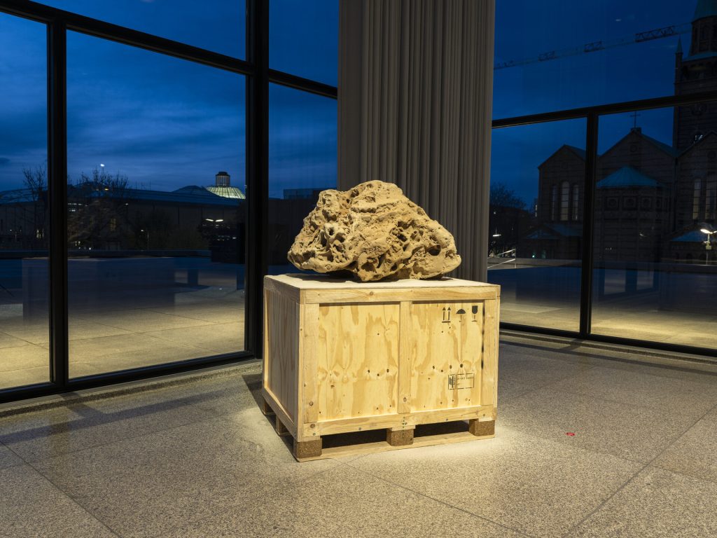 A 3D-printed sand sculpture sits on a wooden crate in front of a large window.