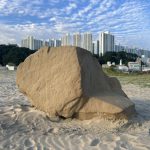 A large sand sculpture sits on the beach with city buildings in the background.