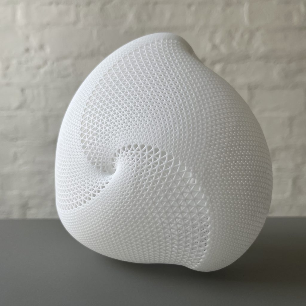 A white, 3D-printed resin sphere with a geometric lattice pattern sits on a gray surface against a brick wall.