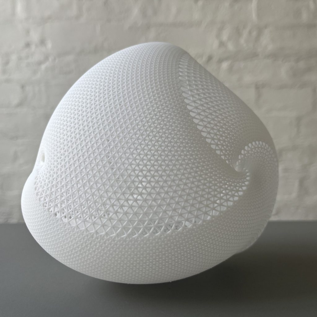 A white, 3D-printed resin sphere with a lattice pattern sits on a gray surface against a white brick wall.