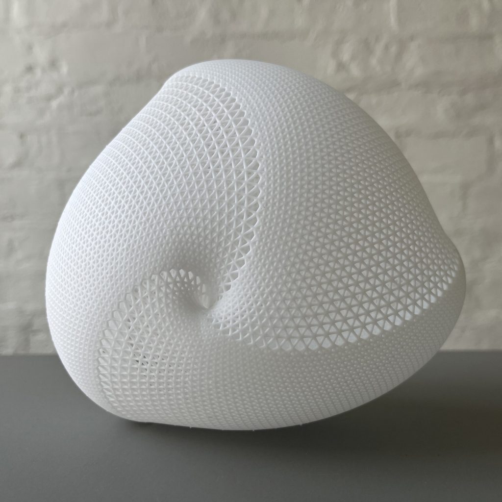 A white, 3D-printed resin object with a geometric lattice pattern sits on a gray surface against a brick wall.