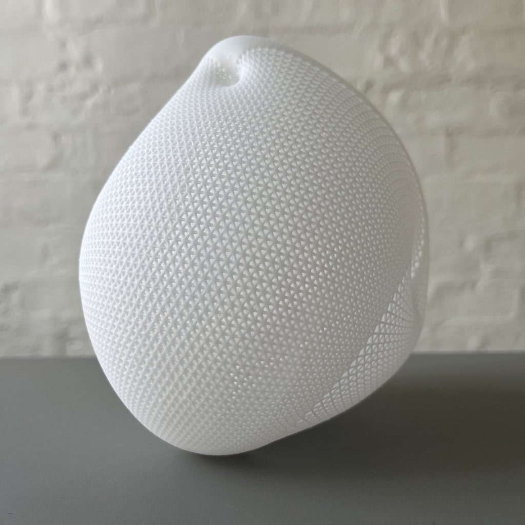 A white, 3D-printed resin object with a grid pattern sits on a gray surface against a brick wall.