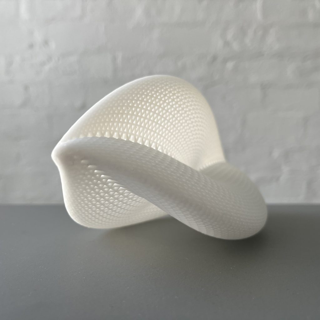 A white, 3D-printed resin hypergraph structure with a textured surface sits on a gray surface against a white brick wall.