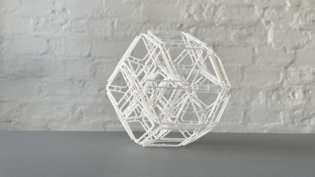 A white 3D-printed resin structure composed of interconnected rhombic shapes sits on a gray surface against a white brick wall.